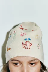 Gorro tipo beanie de punto color crema, con detalle de múltiples bordados coloridos que incluyen figuras como un perro salchicha, flores, lazos y objetos decorativos.