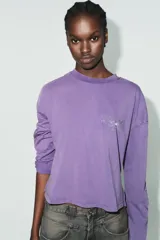 Camiseta de manga larga con corte oversize, confeccionada en algodón color violeta con efecto lavado. Presenta cuello redondo, bajo asimétrico y detalles de texto con aplicación de brillos en el pecho y las mangas.