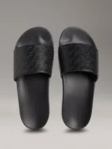 Ojotas tipo slide de color negro con tira superior de piel sintética que presenta un patrón grabado del logo de Calvin Klein. Poseen plantilla ergonómica moldeada y suela de EVA ligera.