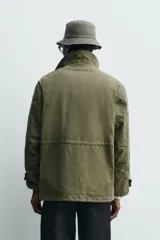 Cazadora de estilo militar en color verde oliva, confeccionada en algodón con efecto lavado. Presenta un diseño de corte relajado con cuello doble, cierre frontal con cremallera oculta por solapa con botones a presión, cuatro bolsillos frontales de plastrón con solapa y puños ajustables con botón.