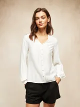 Blusa de color blanco hueso con escote en V y cierre frontal de botones pequeños. Presenta mangas largas con puños ajustados y un diseño de corte recto.