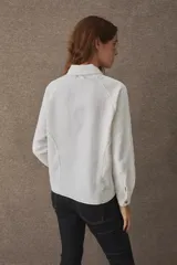 T-shirt de manga larga confeccionada en mezcla de algodón y lino, color blanco, con cuello camisero, cierre frontal parcial con cremallera, puños abotonados y aberturas laterales en el bajo. Cuenta con un pequeño bordado distintivo en el pecho.
