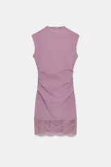 Vestido corto de tejido elástico color lila, con cuello redondo, diseño sin mangas y detalle de frunces laterales en la cintura. El bajo presenta una terminación con encaje combinado a tono.