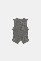 Chaleco de tejido texturado en tonos claros (blanco/beige/gris) con escote pico y cierre frontal de cuatro botones dorados. Presenta un bajo asimétrico.