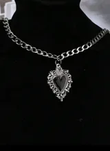 Collar plateado de cadena gruesa con dije de corazón negro con marco ornamentado y detalle de llama en la parte superior.