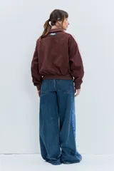 Cazadora de estilo deportivo en color marrón, confeccionada en tejido tipo gamuza. Presenta cierre frontal completo, cuello alto, puños y ruedo acanalados, y franjas laterales en tono rosado. Incluye el logo de la marca bordado en el pecho.