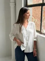 Blusa blanca de manga larga con cuello en V y cierre frontal de botones. Presenta una textura sutil en la tela y un corte recto y holgado.