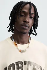 Camiseta de manga corta color crema, corte boxy fit, con cuello redondo. Presenta un estampado frontal en negro con el texto 'VORTEX PULSE' y el número '1974' en un óvalo rojo desgastado. Tiene pequeños detalles gráficos rojos en las mangas.