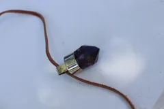 Collar corto con dije de cuarzo en punta de color violeta oscuro, montado en una base metálica plateada y suspendido de un tiento de cuero marrón.