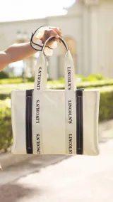 Bolso tipo tote bag color crudo, con asas de mano y detalles en símil cuero color marrón. Tiene estampado vertical con la inscripción "Lincoln's".