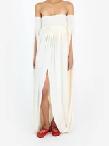 Vestido largo color blanco de silueta fluida, con escote palabra de honor, cuerpo fruncido tipo smock y tajo frontal. Presenta mangas largas desmontadas con aberturas y diseño abullonado en los brazos.