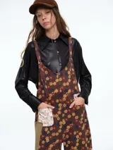 Mono de corte holgado con estampado floral sobre fondo marrón. Presenta tirantes ajustables, bolsillos frontales con parches en contraste y diseño de pierna ancha.