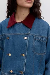 Cazadora de denim azul con corte cropped y diseño de doble botonadura frontal. Presenta un cuello de pana en color bordó y botones metálicos dorados.