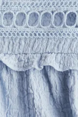 Blusa celeste de manga larga con textura arrugada, cuello alto con volados y abertura frontal con lazos para atar. Presenta canesú con volados y detalles de encaje calado en el pecho y hombros. Los puños terminan con elástico y volado.