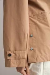 Trench color camel con capucha desmontable, cierre frontal con cremallera, bolsillos laterales y mangas largas con ajuste en puños.