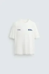 Camiseta de manga corta y cuello redondo, color blanco roto, con efecto de doble capa (la capa inferior gris asoma en el ruedo). Presenta estampados pequeños en azul marino en el pecho: un logo con texto 'Idle Eden' y otros símbolos.