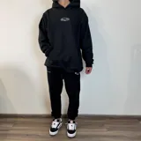Canguro negro de corte oversize con capucha y bolsillo frontal tipo canguro. Presenta un estampado ovalado blanco en el pecho con la inscripción 'Buena Vibra' y un detalle bordado en el puño de la manga.