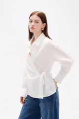 Camisa blanca de algodón con diseño cruzado y anudado lateral, cuello camisero y mangas largas con puños.