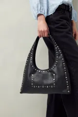 Cartera de hombro en símil cuero negro con apliques de tachas metálicas plateadas que recorren el contorno y el bolsillo frontal. Presenta una silueta curva y diseño funcional.