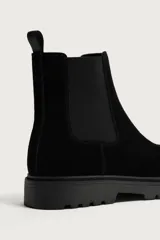 Bota tipo Chelsea de hombre en color negro, confeccionada en cuero con acabado de serraje. Cuenta con elásticos laterales, tirador trasero y suela dentada con volumen de estilo track.
