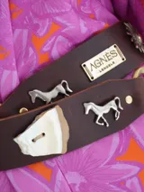 Cinto ancho de cuero marrón con apliques metálicos decorativos en forma de sol, caballo y herraduras, acompañado de tachas.