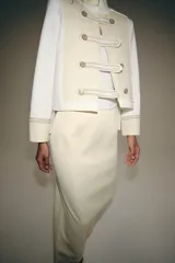 Chaqueta de estilo militar de ZARA Woman Collection, confeccionada en mezcla de algodón en color blanco y beige. Presenta cuello subido, manga larga con puños anchos en contraste y cierre frontal con alamares y botones metálicos en relieve. Tiene bajo asimétrico con abertura en la espalda.
