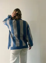 Sweater de punto con diseño de rayas verticales en tonos azul y gris, con sutil acabado brillante. Presenta cuello redondo, mangas largas con puños ajustados y corte amplio con aberturas laterales en el ruedo.
