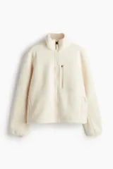 Campera deportiva color crema en tejido sherpa, de corte boxy, con cuello elevado, cierre frontal, bolsillo superior con cierre, bolsillos laterales insertados con cierre, elástico fino en los puños y cordón de ajuste en el bajo.
