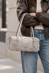 Cartera de cuero vacuno tipo baguette, de color gris claro con acabado vintage y diseño alargado. Cuenta con dos asas largas para llevar al hombro, cierre superior y el logo de la marca grabado en el frente.