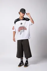 Camiseta blanca de poliéster con cuello en V negro y ribete rojo. Estampado frontal con una estrella negra y texto con efecto desgastado. Mangas cortas con números '0' y '1' en color rojo.