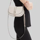 Cartera tipo saddle bag de Guess, color beige, con formato estructurado, solapa frontal con cierre magnético y logo metálico dorado. Incluye correa de hombro con cadena y detalle de tira decorativa.