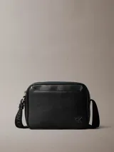 Cartera tipo cámara Calvin Klein de color negro, confeccionada en poliuretano texturizado. Presenta una silueta compacta con cierre de cremallera superior, correa de hombro ajustable con el logo de la marca y monograma CK grabado en la esquina frontal.