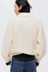 Buzo de punto acanalado color beige, con cuello polo y tapeta frontal con botones metálicos decorativos. Presenta hombros caídos, mangas largas con puños ajustados y diseño de corte recto.