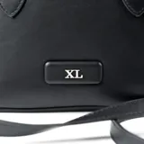 Cartera tipo bowling de la marca XL, confeccionada en símil cuero color negro. Presenta asas fijas, correa regulable extraíble, cierre superior y placa metálica con logo en el frente.