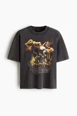 Remera de algodón de punto pesado, color negro con efecto lavado, cuello redondo acanalado y mangas cortas con hombros caídos. Presenta un estampado frontal con un águila, cactus y la inscripción 'Rodeo Sunset'.