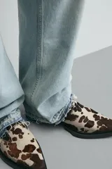 Zueco de piel con pelo, con estampado animal print tipo vaca en tonos marrones y beige. Presenta una suela de goma a contraste con un ligero tacón cuadrado y punta redondeada.