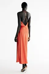 Vestido midi satinado de color naranja tostado, con escote fluido tipo cowl y tirantes finos cruzados en la espalda con detalle de aplicación metálica. Presenta un diseño largo con tajo lateral en la falda.