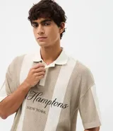 Remera polo de punto con diseño de rayas verticales en tonos beige y blanco. Presenta un corte oversized, cuello clásico con abertura de botones y bordado frontal con la inscripción The Hamptons.