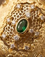 Anillo ancho de bronce con baño de oro, diseño texturizado con detalles calados tipo filigrana, incrustaciones de circonitas y una piedra verde central ovalada.