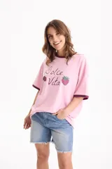 Remera de algodón rosada con diseño oversize y mangas cortas de corte loose fit. Presenta un estampado frontal con la frase "Dolce Vita" y dibujos de fresas, además de ribetes en contraste de tono oscuro en el cuello y los puños.