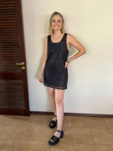 Vestido corto negro de lentejuelas, sin mangas y con escote redondeado. Presenta un ruedo con detalle de tela de red o tul transparente.
