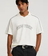 Remera tipo polo de hombre, color blanco crudo, con escote en V ribeteado en negro y ribetes negros en las costuras de los hombros. Presenta la inscripción arqueada "NEW YORK" en negro en el pecho.
