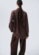 Camisa de seda de corte oversize, color marrón chocolate, con cuello camisero, mangas largas con puños abotonados y cierre frontal de botones ocultos.