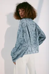 Camisa azul con cuello de solapa, escote en pico y mangas largas con puños abotonados. Presenta un diseño con bordados perforados y patrones geométricos y florales. Cierre frontal con botones.