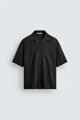 Camisa negra de manga corta con cuello bowling y corte relajado (relaxed fit). Presenta un bolsillo de parche en el pecho y cierre frontal de botones.