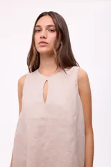 Vestido corto beige de lino, sin mangas, con escote redondo y detalle de abertura en forma de lágrima en el pecho. Presenta diseño holgado y dobladillo asimétrico más largo en la parte trasera con volados.