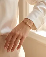 Pulsera tipo esclava rígida de acero con baño de oro, con un diseño minimalista que presenta un detalle de nudo central.