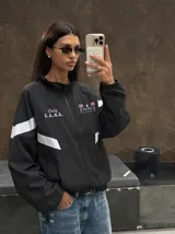 Campera deportiva negra con cuello alto, cierre frontal completo y franjas blancas en las mangas. Presenta estampados en color rosado en el pecho, incluyendo la inscripción "Only F.A.N.S." y gráficos de palmeras con estrellas.