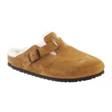 Zueco tipo mule de gamuza color tostado, con interior forrado en piel sintética blanca. Presenta una tira ajustable con hebilla metálica en el empeine y suela de goma con textura.