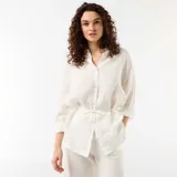 Camisa larga blanca de lino, con cierre frontal de botones, mangas largas y lazo ajustable en la cintura.